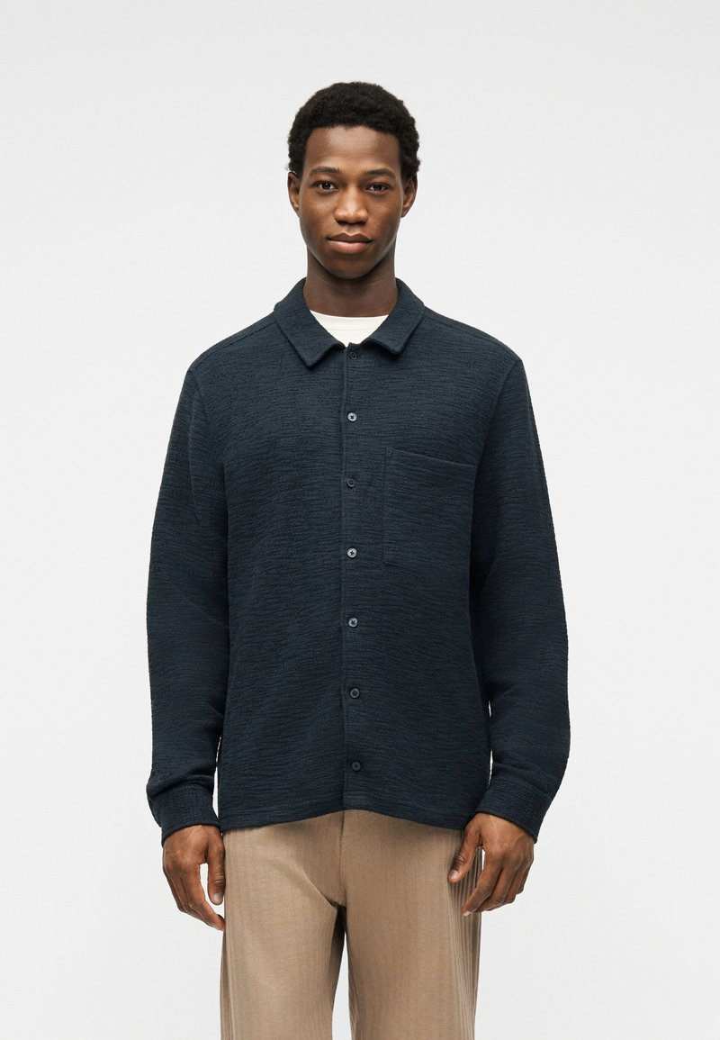 Jeune homme portant une chemise à manches longues texturée de couleur foncée sur un t-shirt blanc, avec un pantalon beige, debout devant un fond clair uni.