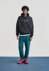 Zwarte hoodie met grijs logo, teal sweatpants en roze sportschoenen. Model staat op een blauw tapijt met een effen achtergrond.
