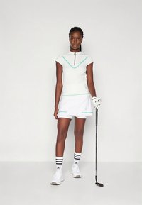 Golf öltözék fehér, rövid ujjú cipzáras felsővel, mely mentazöld részletekkel díszített, és hozzáillő fehér szoknyával. Készlet kiegészítve csíkos zoknival és golfütővel.