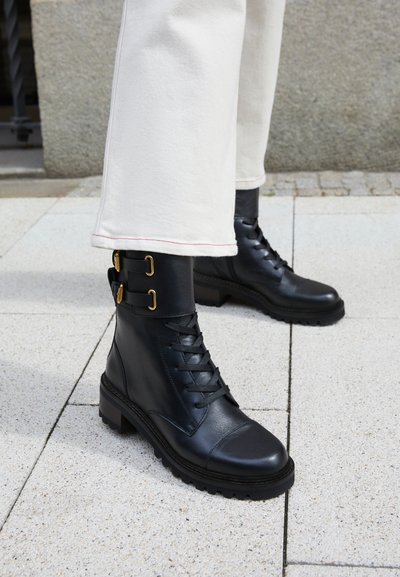 Black leather combat boots with lace-up front, gold accent buckles, and a textured sole. Paired with light-colored, tailored trousers.