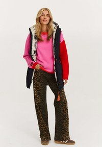 Frau trägt einen rosa und roten Pullover, eine schwarze gesteppte Weste mit Pelzfutter, eine Hose mit Leopardenmuster und braune Sneakers, steht mit einer Hand in der Tasche.