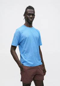 Helehall sinine puuvillane T-särk lühikeste varrukatega, ümmarguse kaelusega, pruunide lühikeste pükstega. Ühtlane tekstuur, lihtne disain ilma mustriteta.