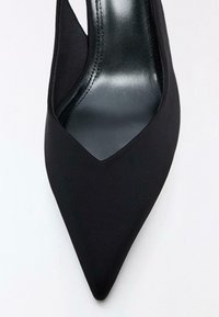 Pantof negru cu vârf ascuțit, partea superioară din material textil, laterale decupate și o talpă interioară netedă, lucioasă, de culoare neagră. Designul include cusături discrete.