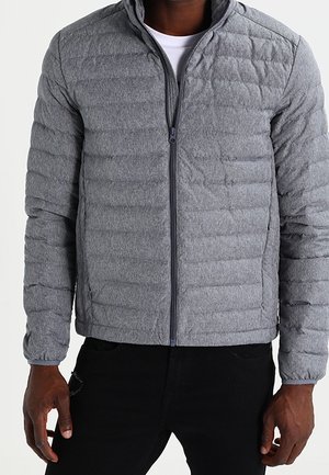 Homme portant une veste matelassée grise avec fermeture éclair sur une chemise blanche et un pantalon noir, debout les mains détendues le long du corps.