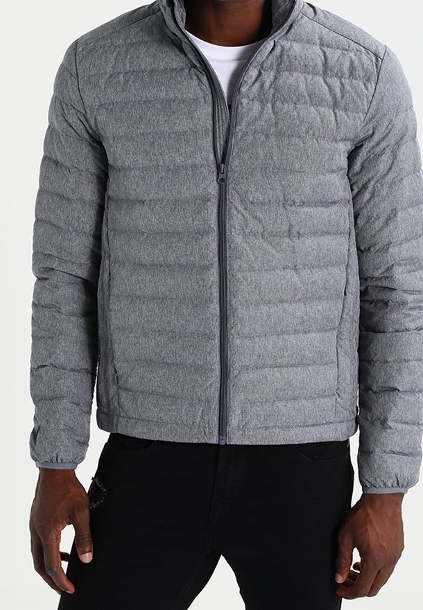 Homme portant une veste matelassée grise avec fermeture éclair sur une chemise blanche et un pantalon noir, debout les mains détendues le long du corps.