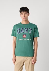 Zelené bavlněné tričko s kulatým výstřihem, které zdobí velký nápis "GANT", logo ve tvaru štítu a bílý nápis "AMERICAN SPORTSWEAR" pod ním.