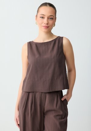 Vrouw met een lichte huid en haar in een staart, draagt gouden oorbellen, een mouwloze bruine top met fijne streepjes en bijpassende hoog getailleerde broek met fijne streepjes, hand in de zak.