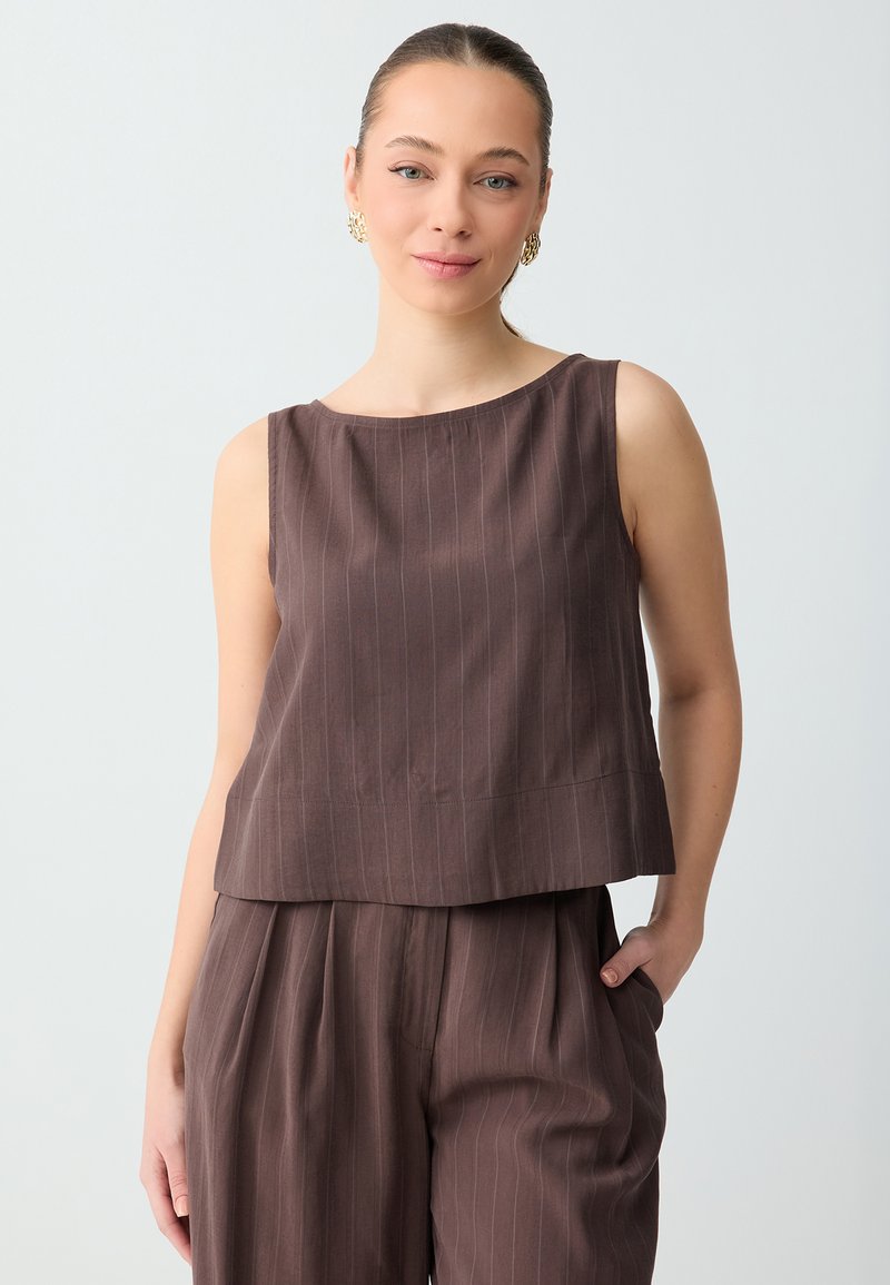 Femme à la peau claire, cheveux attachés, portant des boucles d'oreilles en or, un haut marron sans manches à fines rayures, et un pantalon taille haute assorti à fines rayures, main dans la poche.
