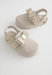 Jasno beżowe zamszowe klapki dla dzieci z regulowanymi złotymi klamrami i tylnymi paskami na białym tle.