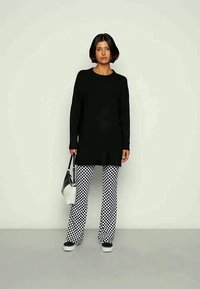 Svart langermet tunika, kombinert med svart-hvite rutete vide bukser. Modellen holder en svart og hvit tote bag og har på seg slip-on sko.