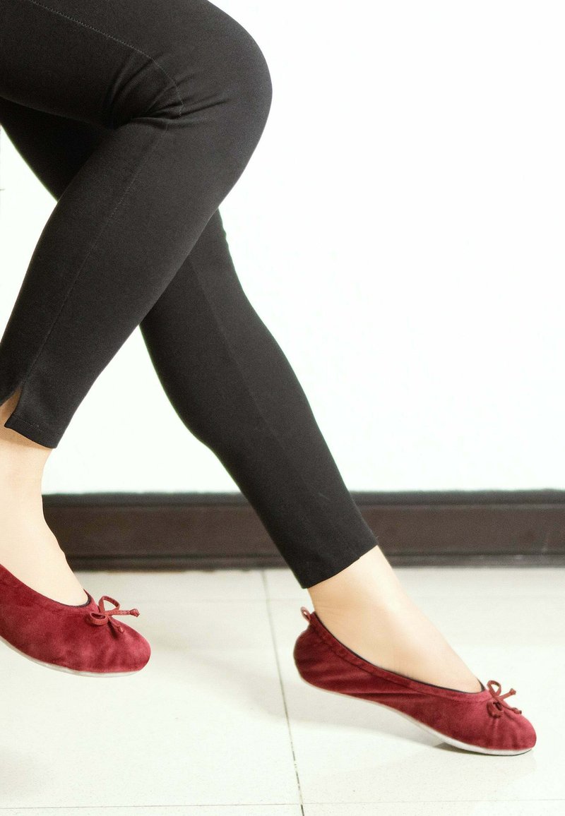 Ballerines en velours bordeaux avec un bout rond et un détail de nœud, associées à des leggings noirs, mettant en avant une texture lisse.