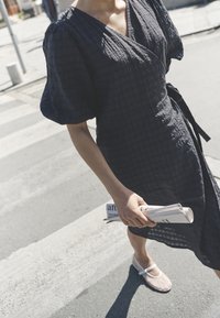 Tamsiai mėlyna aptempta suknelė su tekstūruotu audiniu, trumpomis pūstomis rankovėmis ir surištu liemeniu. Asmuo laiko sulankstytas laikraščio puslapius ir dėvi lengvas, plokščias batus.