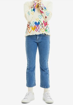 Persona con vaqueros azules cortos, zapatillas blancas y un suéter crema decorado con patrones de corazones de colores, manos cubriendo el rostro.