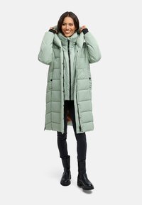 Lichtgroene pufferjas met een capuchon, ritssluiting en zwarte accenten. Heeft een lange, gequiltte stijl en twee voorzakken. Zwarte broek en laarzen.