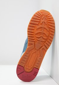 Semelle d'une chaussure New Balance bleue avec une bande de caoutchouc orange et rouge, affichant le nom de la marque et le texte "XAR 1000".