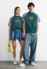 Νεαρή γυναίκα και άνδρας φορούνται ταιριαστά πράσινα μπλουζάκια με σχέδια, φούστα από τζιν και τζιν παντελόνι, λευκά αθλητικά παπούτσια· η γυναίκα κρατά μια μικρή κίτρινη τσάντα χειρός.