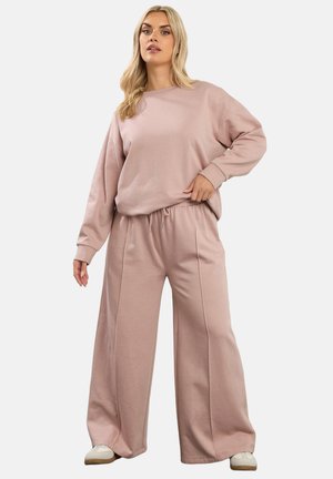 Blondynka w luźnej, pudrowo-różowej bluzie i szerokich spodniach z szerokimi nogawkami oraz białych trampkach, stojąca na jednolitym tle.