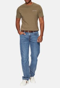 T-shirt verde oliva a maniche corte con logo 'Guess Jeans', abbinato a jeans in denim blu e sneakers bianche, con vestibilità standard.