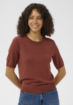 Femme aux cheveux bouclés souriante, portant un pull à manches courtes de couleur rouille et un jean bleu, debout devant un fond uni.