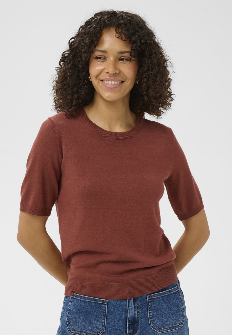 Femme aux cheveux bouclés souriante, portant un pull à manches courtes de couleur rouille et un jean bleu, debout devant un fond uni.