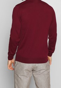 Bordeauxfarbener Pullover mit gerippten Manschetten und Saum, aus einem weichen Strickmaterial gefertigt. Getragen mit hellgrauen, strukturierten Hosen.