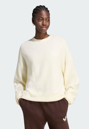 Femme aux cheveux tressés portant un pull crème et un pantalon marron, les mains dans les poches, regardant légèrement sur le côté et souriant doucement.