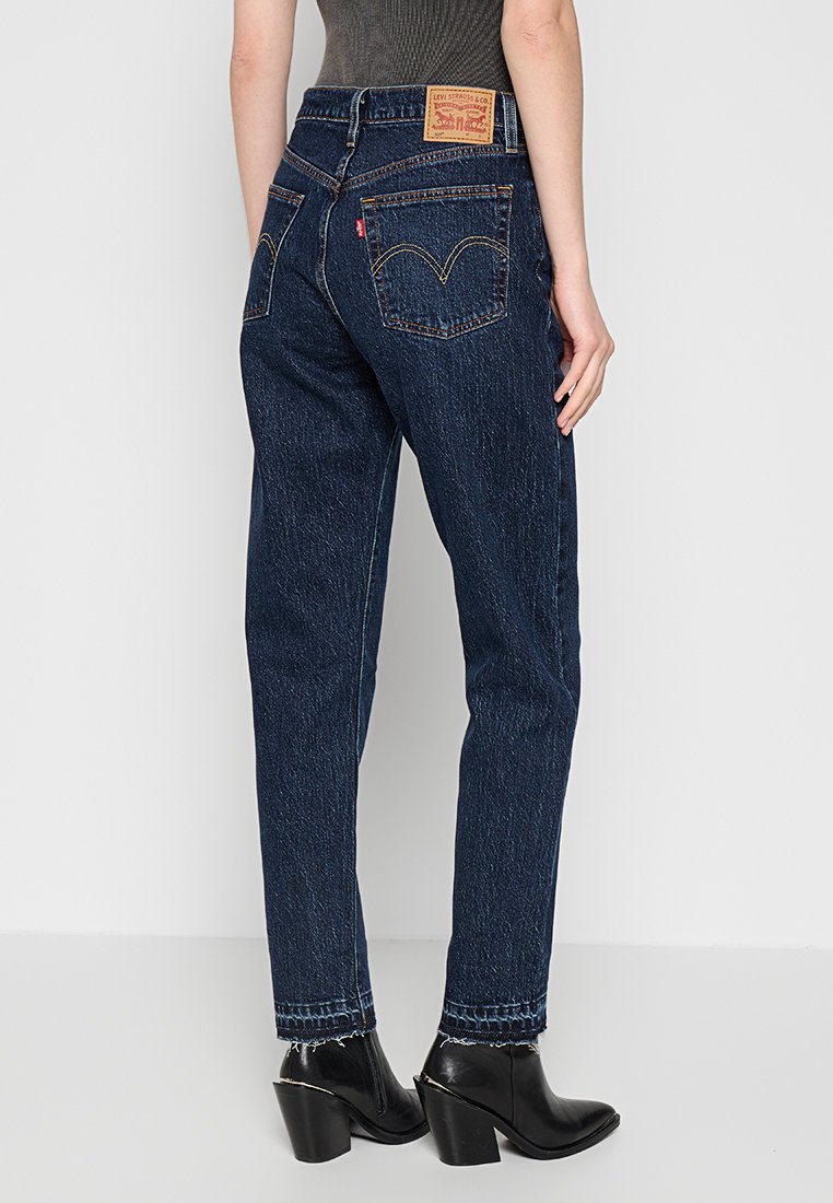 Jean en denim bleu foncé avec une coupe décontractée, arborant une étiquette en cuir à l'arrière de la taille, deux poches arrière et un ourlet effiloché.