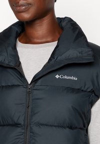 Černá péřová vesta s vysokým límcem, zipovým uzávěrem a logem Columbia na hrudi. Nositá přes světle šedou košili. Texturovaný, zateplený design.
