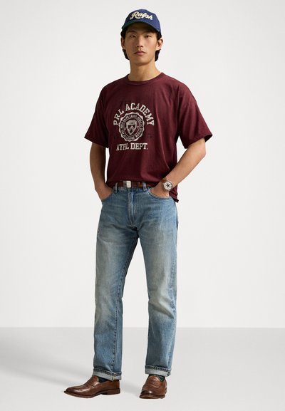 Maroon graphic t-shirt met de tekst "P.R. Academy", lichte spijkerbroek, bruine leren schoenen en een blauwe pet met het "Ralph" logo. Zilveren horloge zichtbaar.