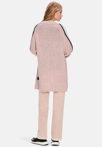 Übergroßer rosa Strickcardigan mit seitlichen Schlitzen, geripptem Saum und kontrastierenden schwarzen Details an den Ärmeln. Kombiniert mit hellbeigen Hosen.