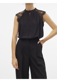 Blouse noire avec manches courtes en dentelle, encolure à ouverture clé et silhouette fluide. Associée à un pantalon noir taille haute.