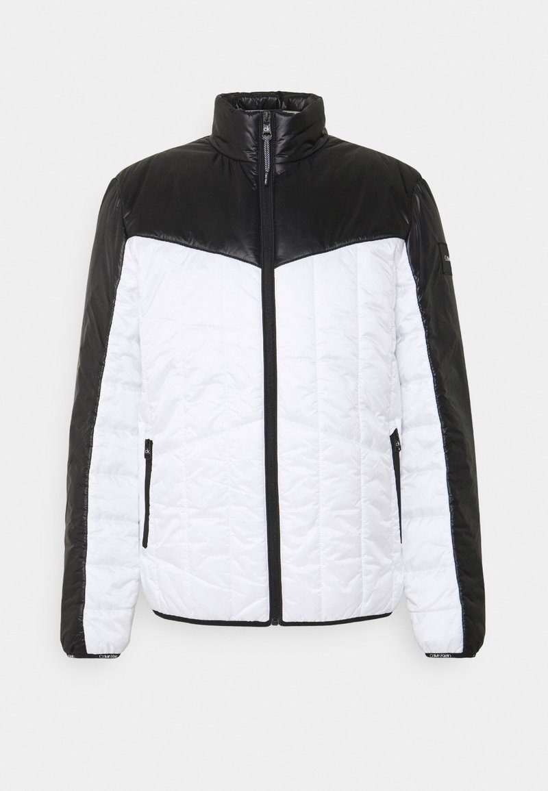 Veste matelassée dotée d'un corps blanc matelassé avec des épaules en similicuir noir, un col montant et une fermeture éclair frontale. Deux poches latérales zippées.