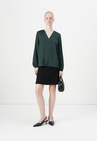 Mørkegrønn bluse med lange ermer og V-hals, kombinert med et svart miniskjørt. Modellen holder en liten svart håndveske og har på seg svarte slingback-hæler.