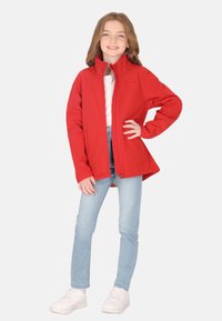Rød softshell-jakke med høj krave, lynlås og sidelommer, båret over en hvid skjorte og lyseblå jeans.