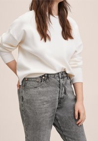 Suéter de punto blanco con puños acanalados, combinado con jeans grises de tiro alto que presentan accesorios de cobre y un ajuste relajado.