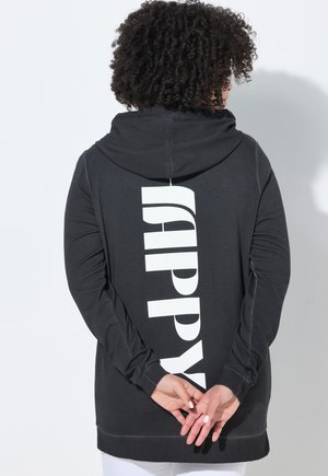 Persona con capelli ricci che indossa una felpa nera con cappuccio con la scritta "HAPPY" in grandi lettere bianche sulla schiena, mani intrecciate dietro.