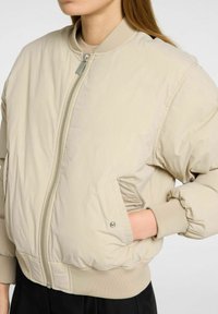 Una donna indossa una giacca bomber beige chiaro con zip e polsini a costine, con le mani in tasca, su uno sfondo neutro.