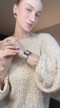 Storas smėlio spalvos megztinis, sidabrinės spalvos tinklelio laikrodis su apvaliu ciferblatu, sluoksniuotos žiedai ir didelės auksinės lankelio formos auskarai. Lygus odos paviršius, matomi tatuiruotės.