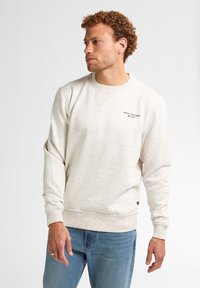 Felpa avorio con motivo marmorizzato, polsini e orlo a coste, scollatura a girocollo e piccolo logo sul petto. Abbinata a jeans azzurri chiari.