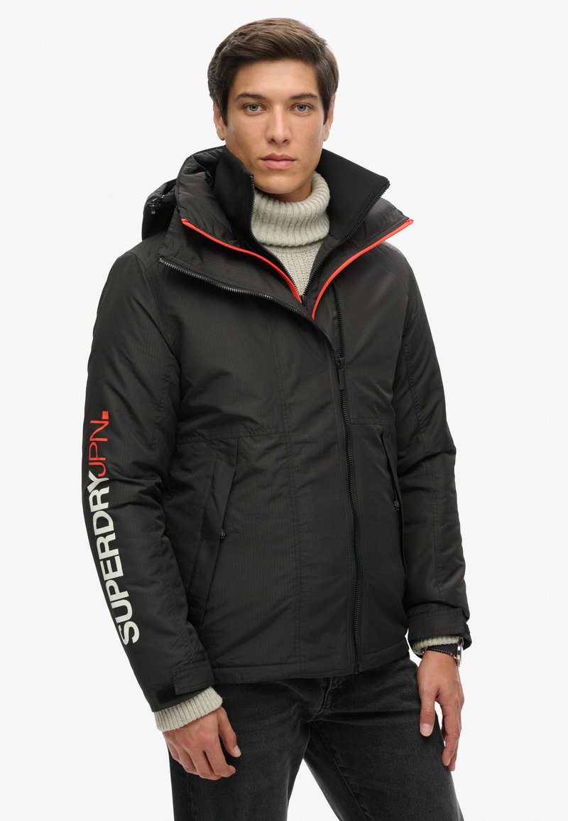 Abrigo acolchado negro con cuello alto, detalles en rojo y un gran logo blanco en la manga. Cuenta con cierre de cremallera en la parte frontal y bolsillos laterales.