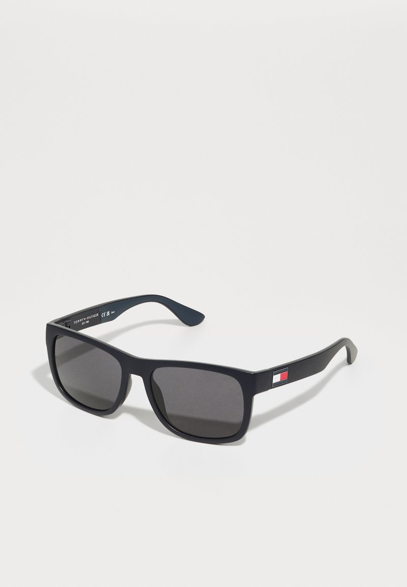 Gafas de sol rectangulares negras con lentes oscuros, que presentan un acento de logo en la patilla. Hechas de plástico con textura suave.