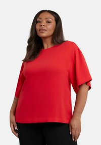 Blusa rossa a maniche corte con scollatura tonda. Tessuto morbido, vestibilità rilassata e design con spalle scese. Abbinata a pantaloni neri.
