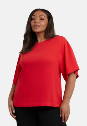 Blusa rossa a maniche corte con scollatura tonda. Tessuto morbido, vestibilità rilassata e design con spalle scese. Abbinata a pantaloni neri.