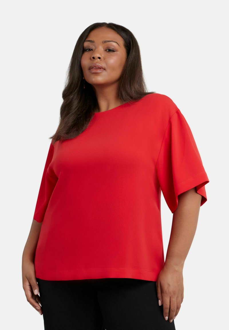 Blusa rossa a maniche corte con scollatura tonda. Tessuto morbido, vestibilità rilassata e design con spalle scese. Abbinata a pantaloni neri.