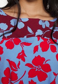 Halenka s květinovým vzorem v modro-burgundské barevné kombinaci. Materiál je lehký s texturovaným vzhledem a velkými červenými květinami.