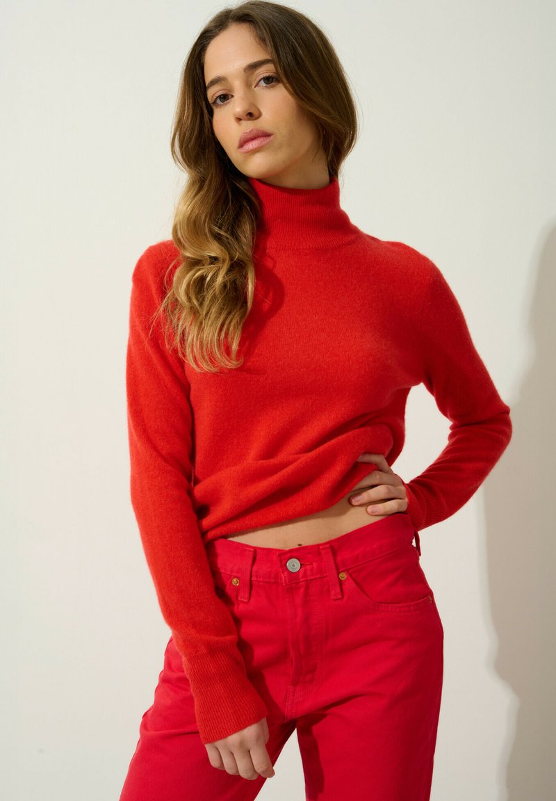 Maglione rosso in lana con collo alto abbinato a jeans rossi a vita alta. Tessuto con trama, maniche lunghe e vestibilità morbida.