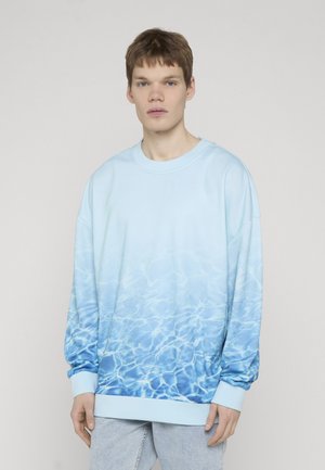 Ung mand iført en lyseblå sweatshirt med et vandbølgemønster, stående foran en ensfarvet lysegrå baggrund.