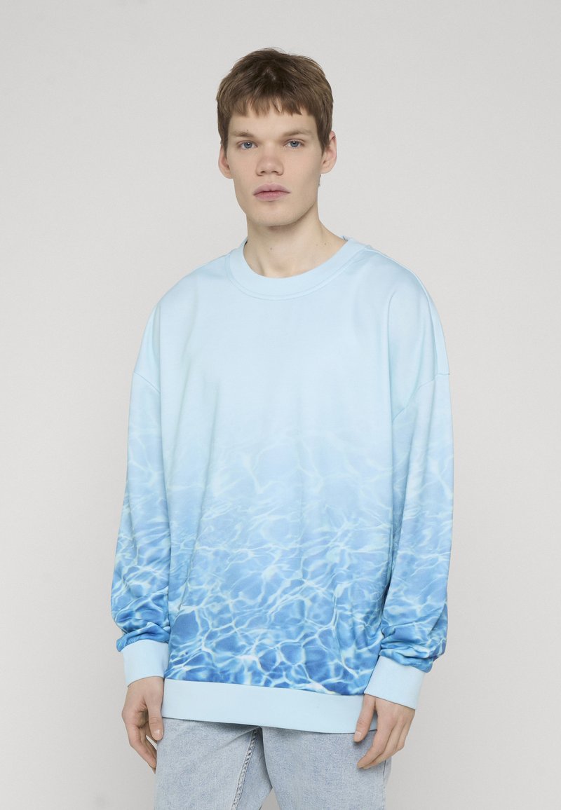 Mladý muž nosiaci svetlomodrú mikinu s motívom vodných vĺn, stojaci pred jednoduchým svetlosivým pozadím.