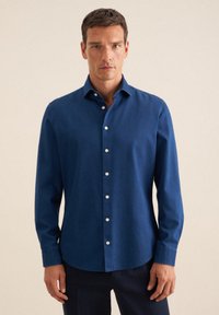 Uomo in piedi rivolto in avanti, che indossa una camicia button-up a maniche lunghe blu scuro con pantaloni neri, su uno sfondo chiaro e uniforme.
