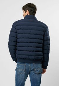 Hombre con chaqueta de quilt azul marino y jeans azules, de espaldas contra un fondo claro y liso.